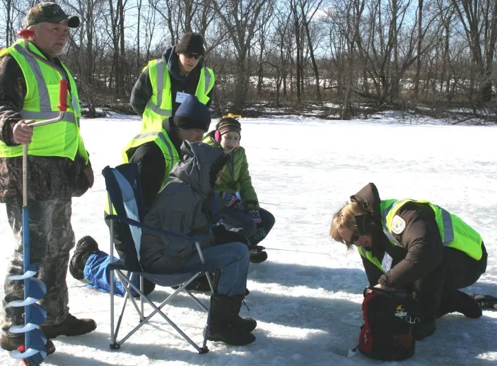 A fishery biologist kneels on the ice and reads a sonar unit at a hole while young anglers fish nearby