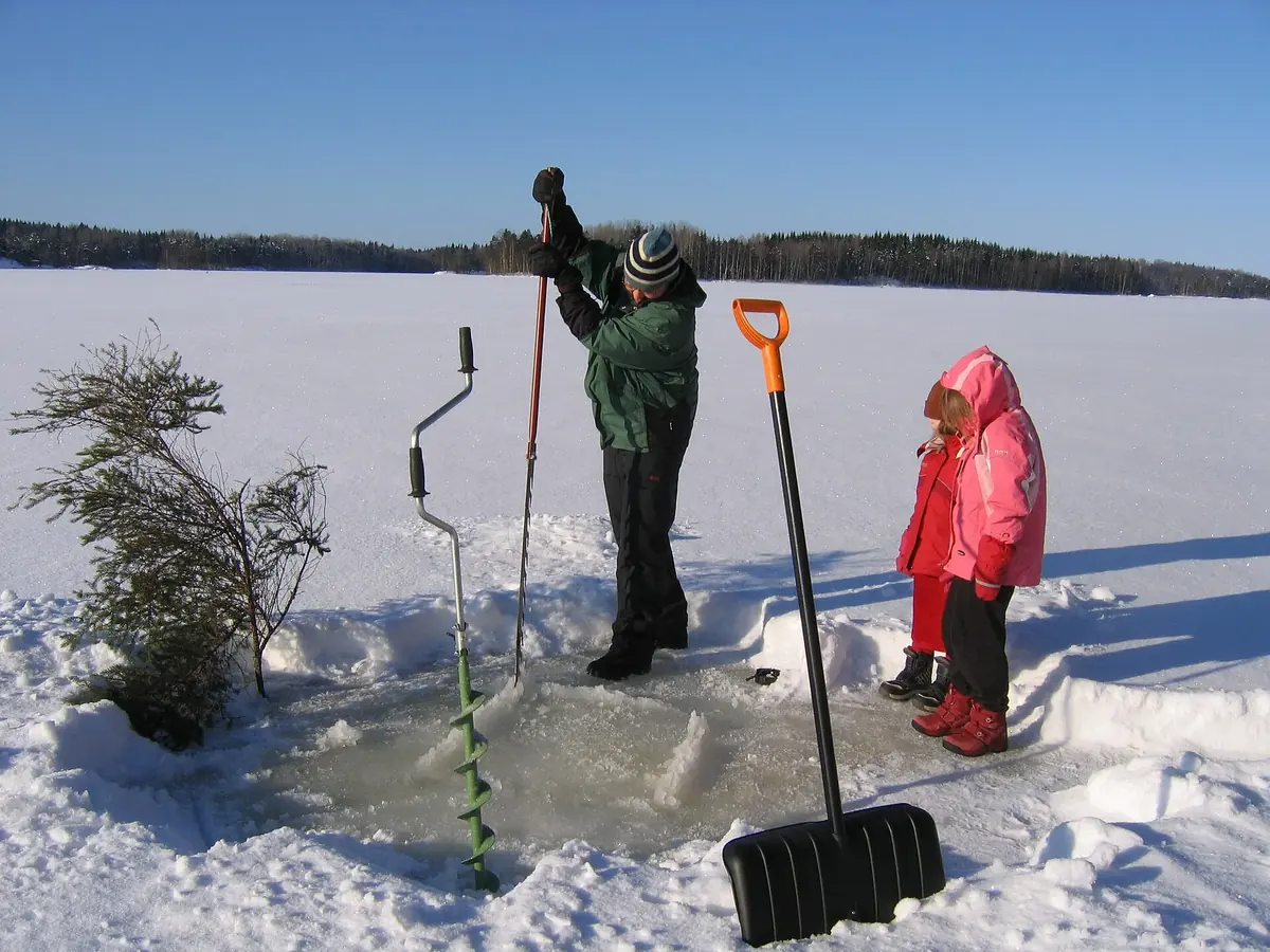 En grön handisborr står kvar i det nyborrade hålet på en frusen sjö, med snötäckt skog på andra sidan.