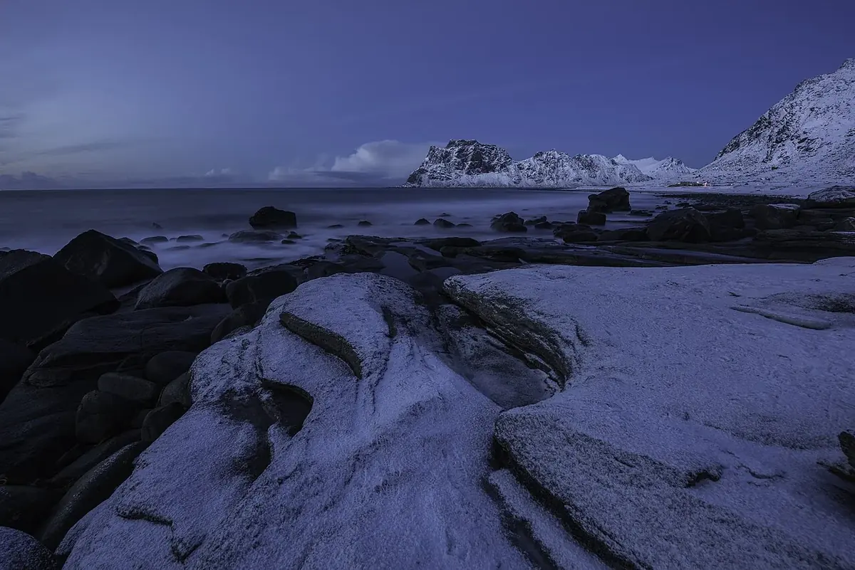 Snødekte svaberg og stein i fjæra ved et vinterhav i Lofoten